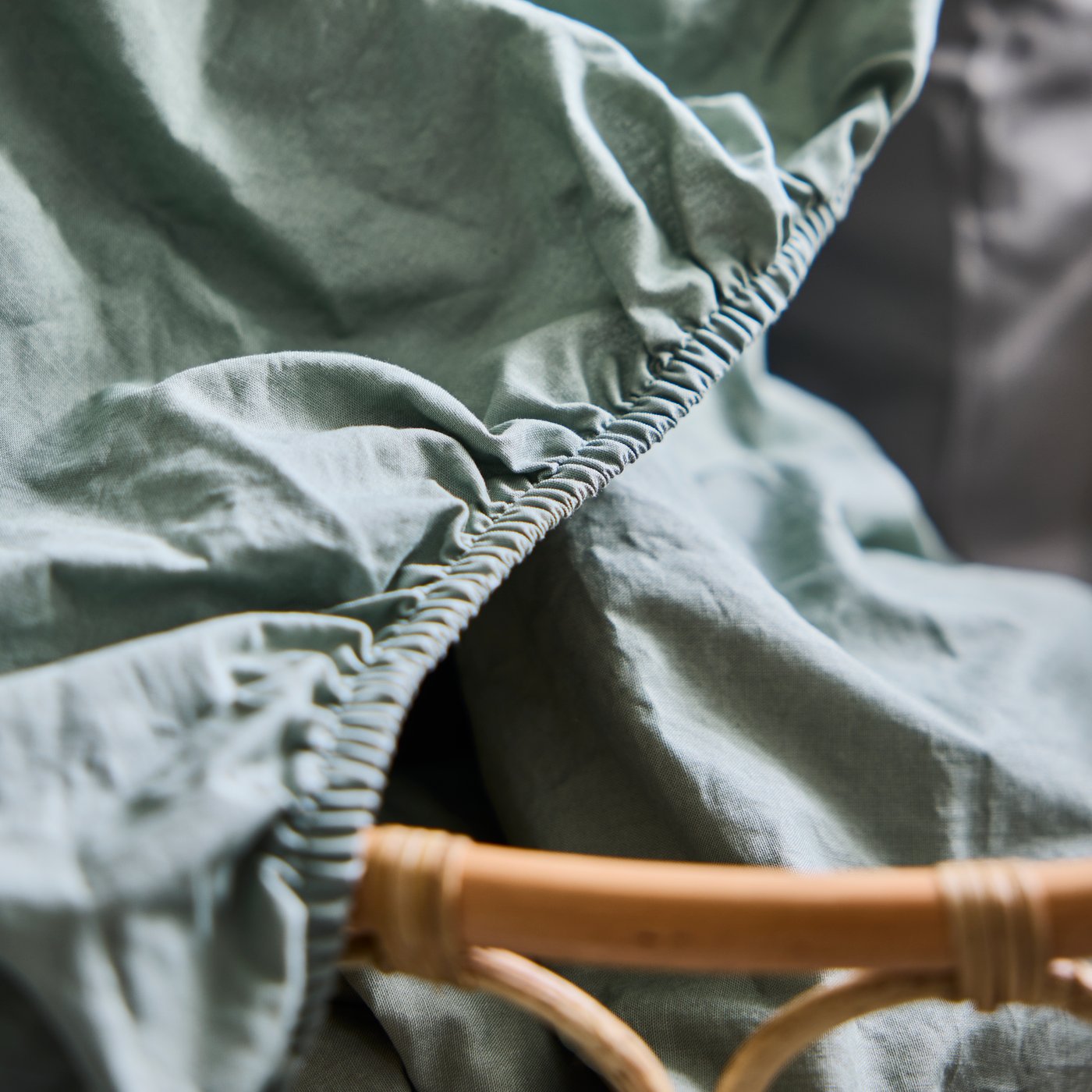 A green DVALA fitted sheet displayed with its elastic edge visible beside a woven basket in a bedroom.