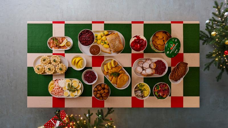 Oversigtsbillede af et julebord dækket med en VINTERFINT dug i røde og grønne tern. På bordet står en masse juleretter på hver deres tallerken.