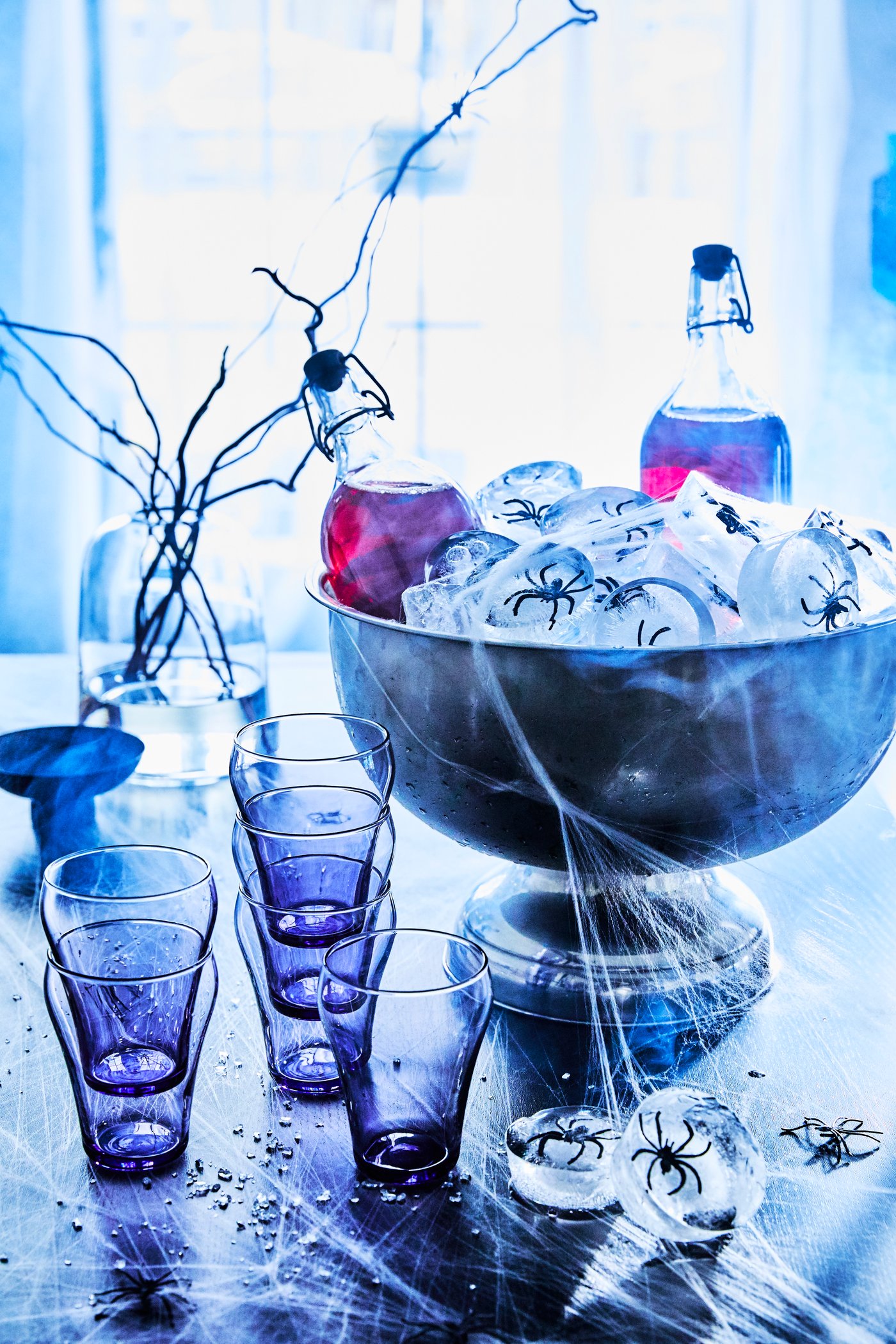A Halloween-inspired table, with Lingonberry drink in KORKEN bottles with stoppers in a large bowl with ice cubes with plastic spiders in them sit on a table with cups and spider webs.