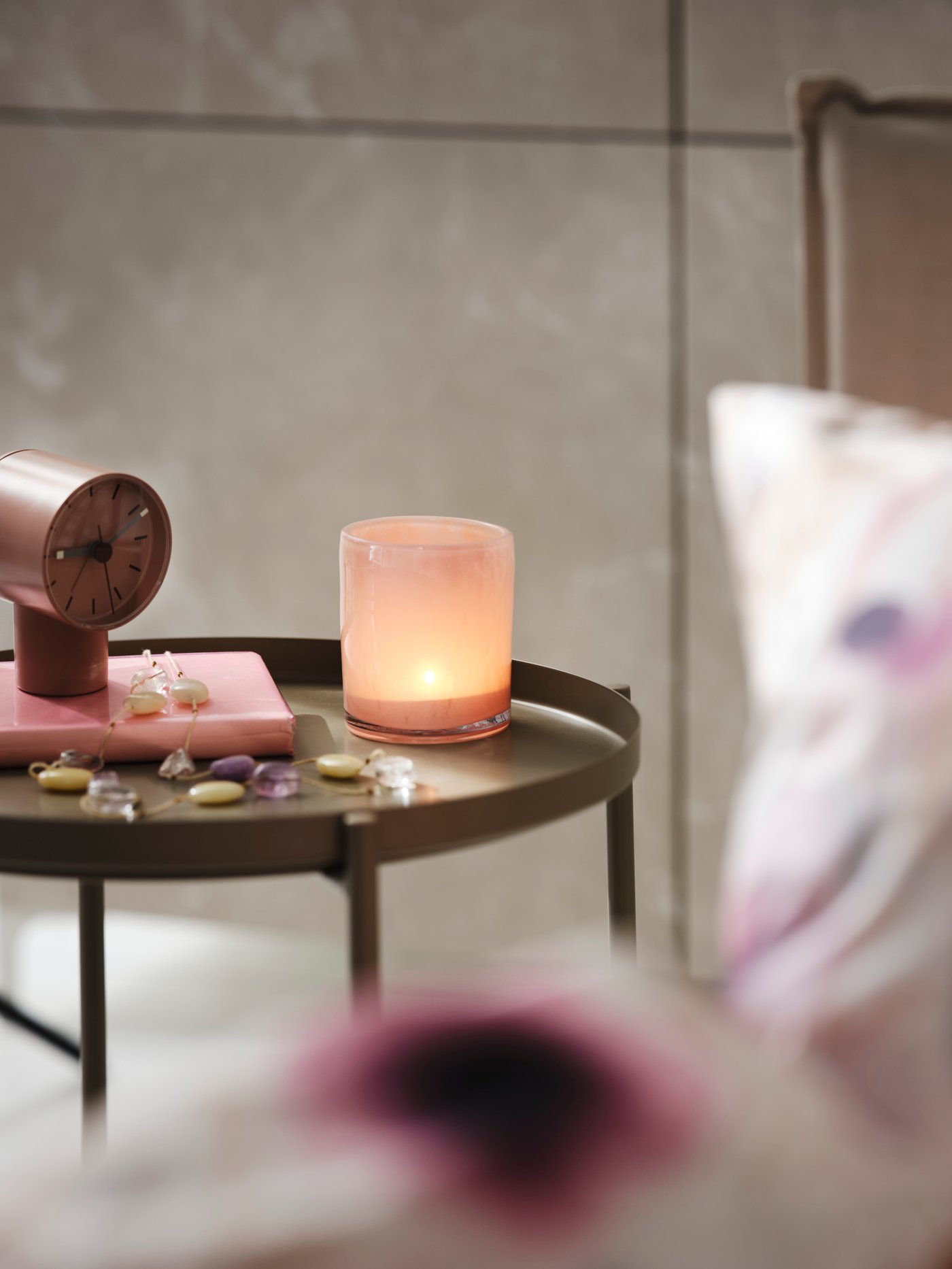 Kulatý odkládací stolek s hořící svíčkou v růžovém skleněném svícnu, vedle růžové hodiny, podložky a drobné dekorace.