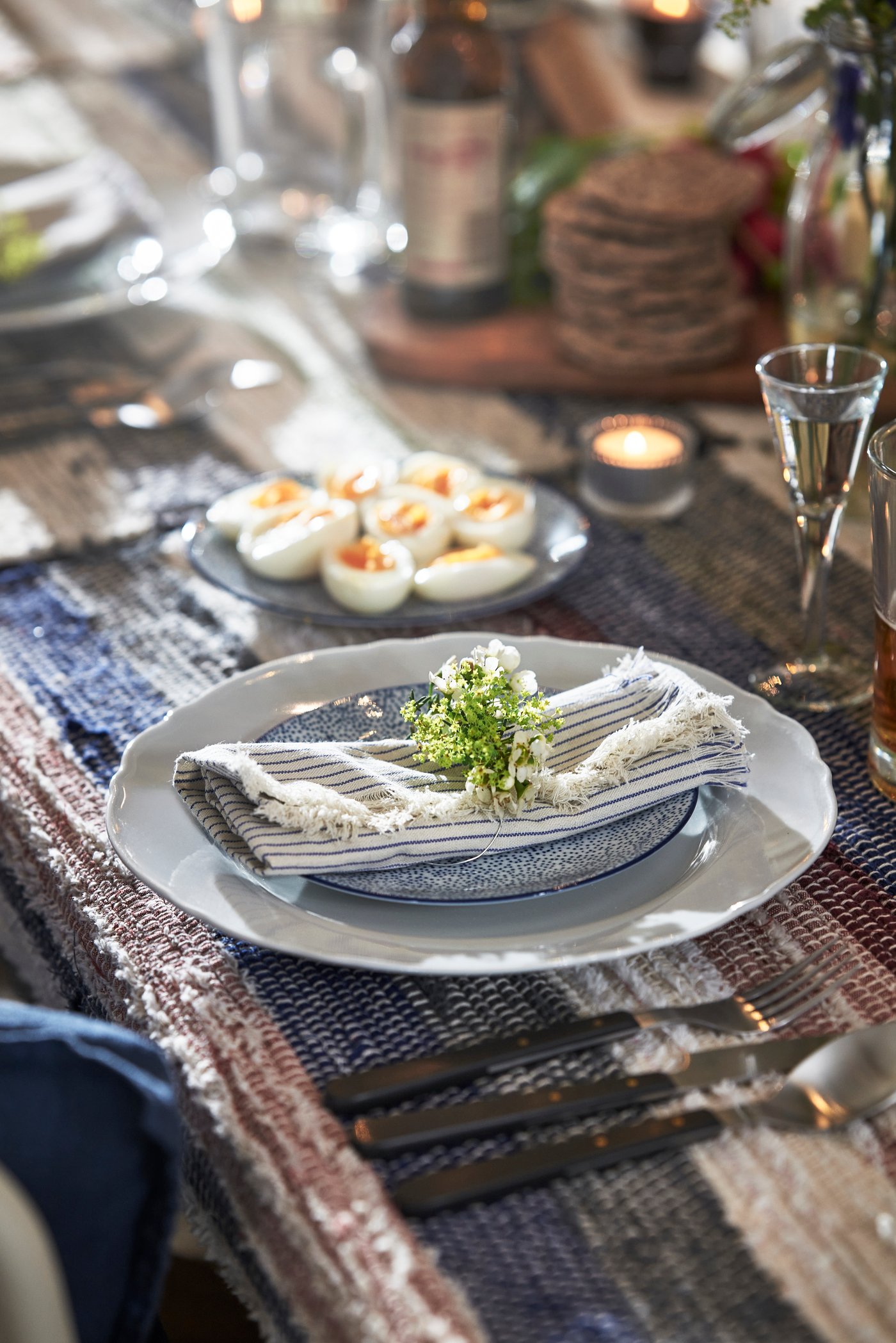 Ett dukat bord med en vit tallrik på mönstrad tallrik och tygservett, toppad med en liten blomdekoration; runtom syns glas, bestick och små förrätter på ett rustikt bord med textilier i naturtoner och mjukt ljus.
