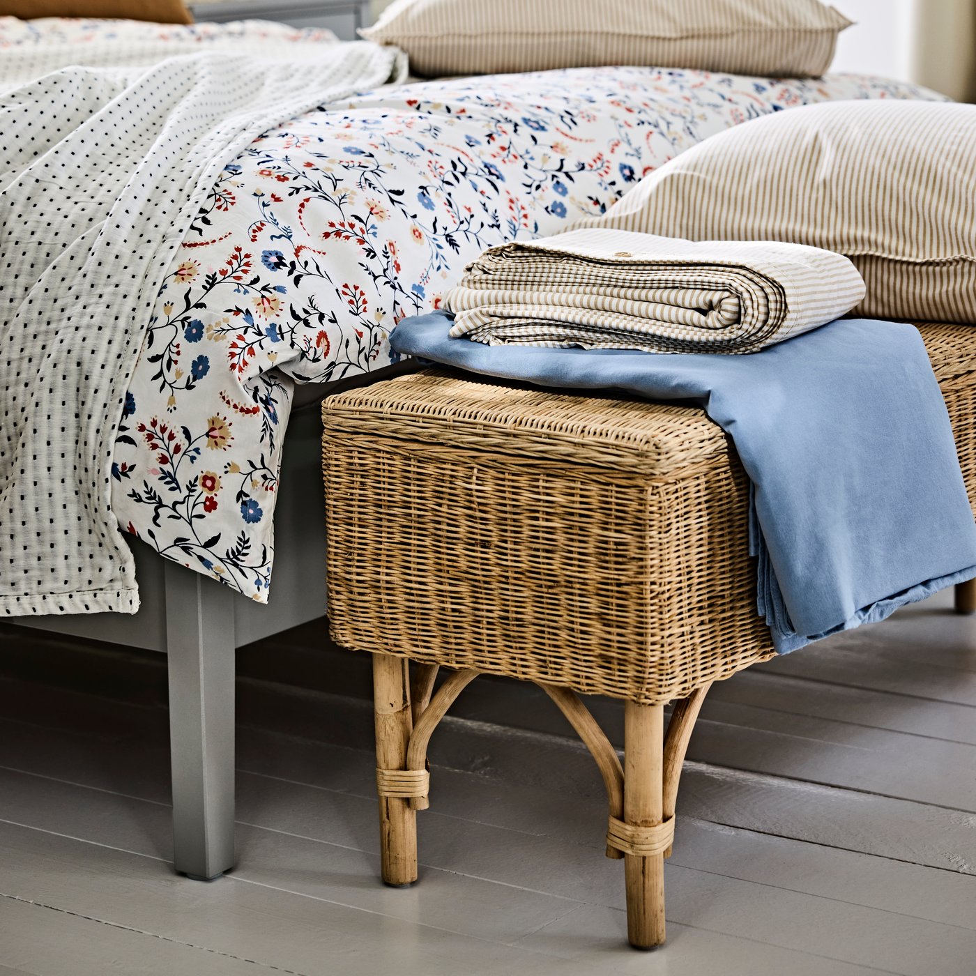 A wicker TOLKNING bench with storage positioned at the foot of the bed and holding folded blankets and bedding.