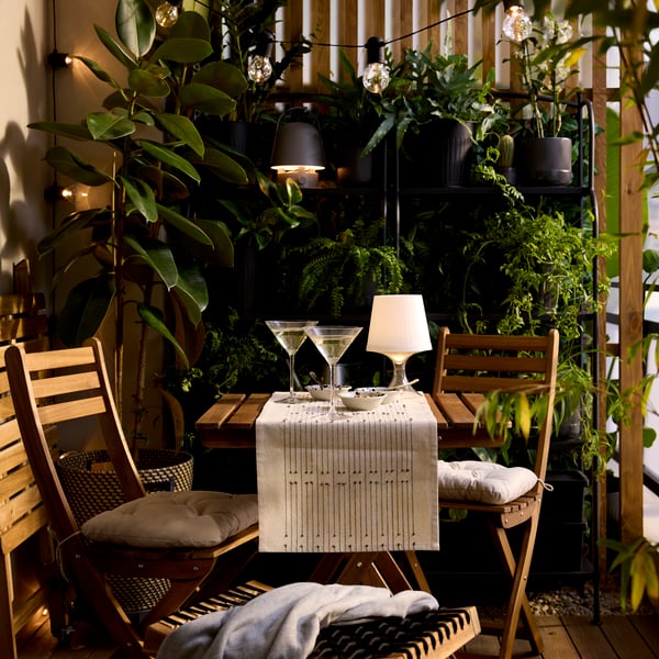 A plastic LAMPAN LED portable lamp with a white shade and clear base is lit, on the table of a bistro set in a lush balcony.