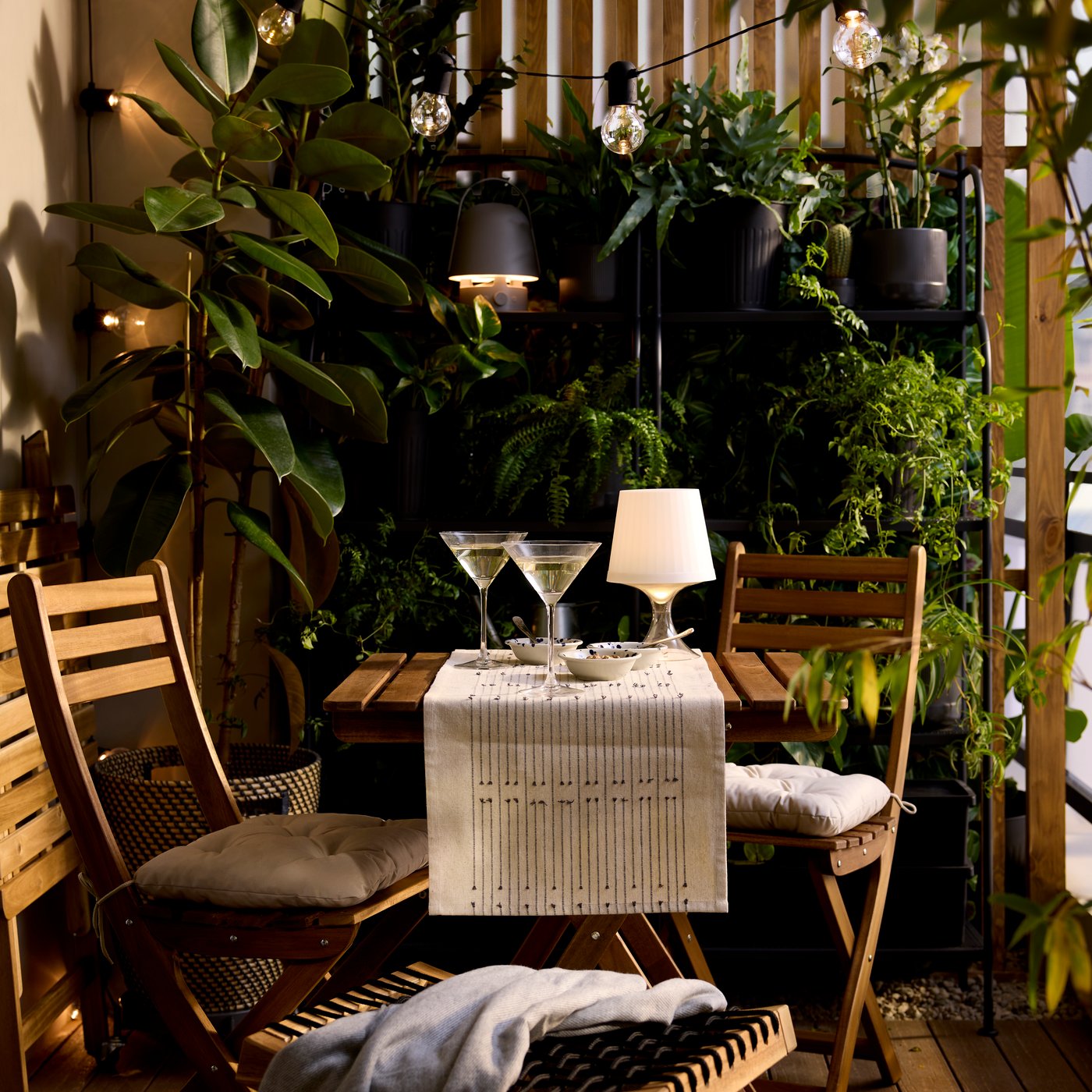 A plastic LAMPAN LED portable lamp with a white shade and clear base is lit, on the table of a bistro set in a lush balcony.