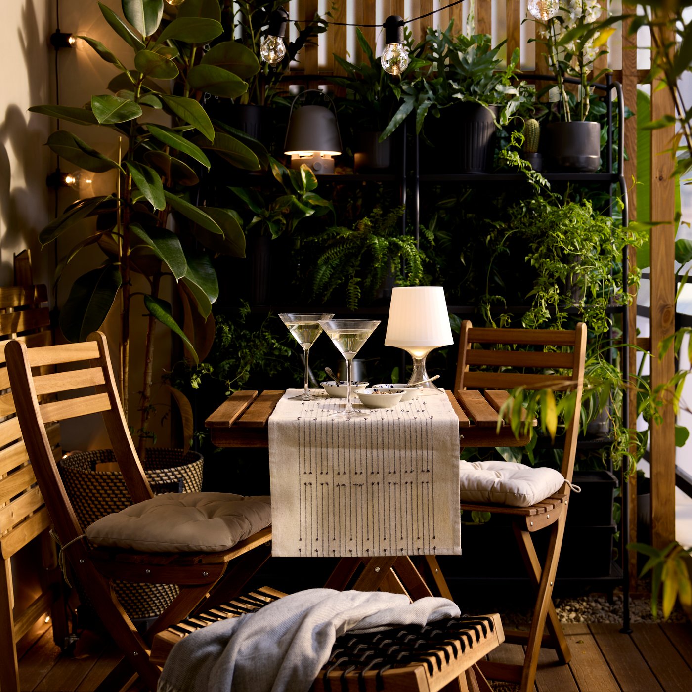 A plastic LAMPAN LED portable lamp with a white shade and clear base is lit, on the table of a bistro set in a lush balcony.