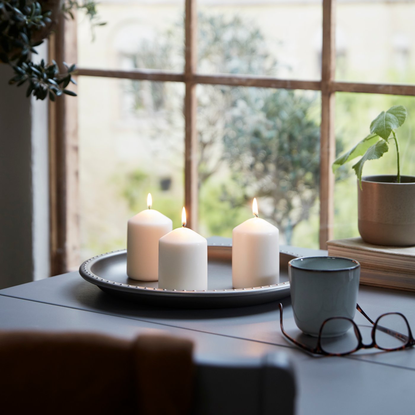 Tři bílé svíčky IKEA na kovovém podnose na stole u okna, vedle hrnek, brýle a květináč s rostlinou.