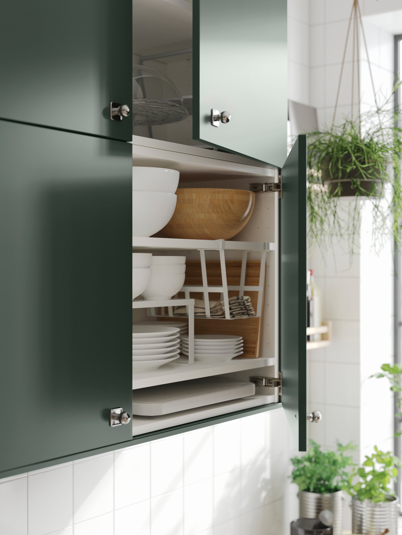 A wall cabinet with deep green kitchen fronts and doors ajar showing plates, bowls, a shelf insert and a clip-on basket.