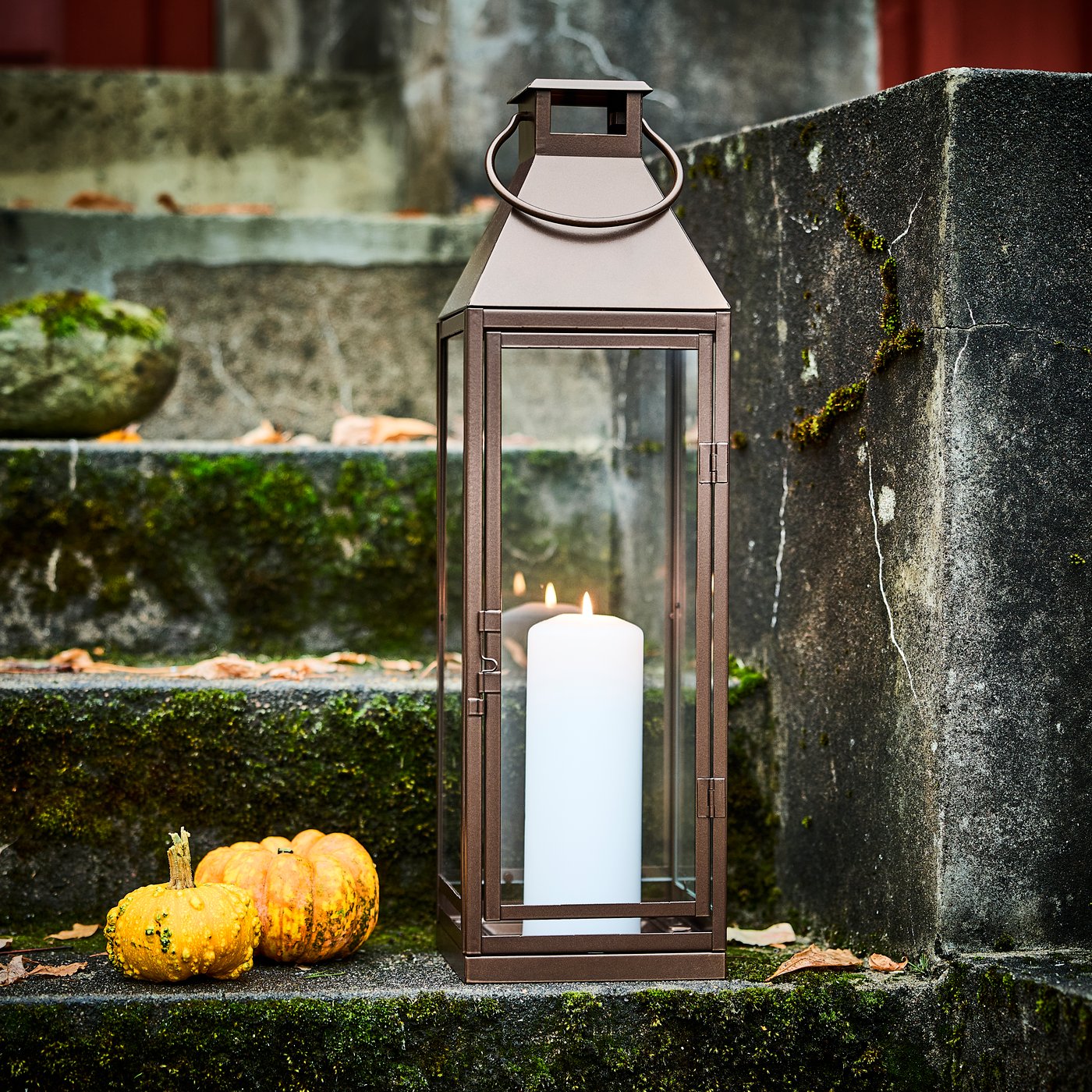 Una vela gruesa blanca encendida dentro de un farol HÖSTAGILLE, junto a dos minicalabazas en una escalera exterior.