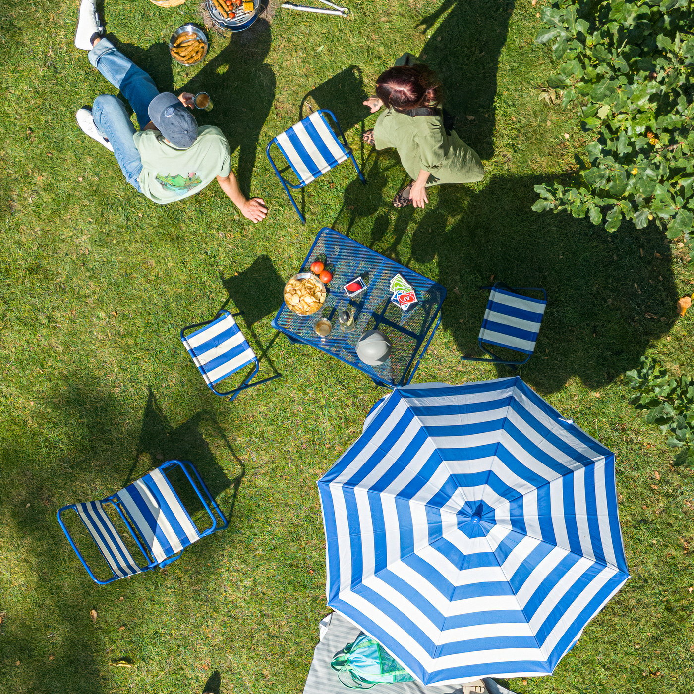 Bild tagen uppifrån på en picknick på gräsmatta med två personer, randiga stolar, STRANDÖN parasoll, snacks, kortspel och grill.