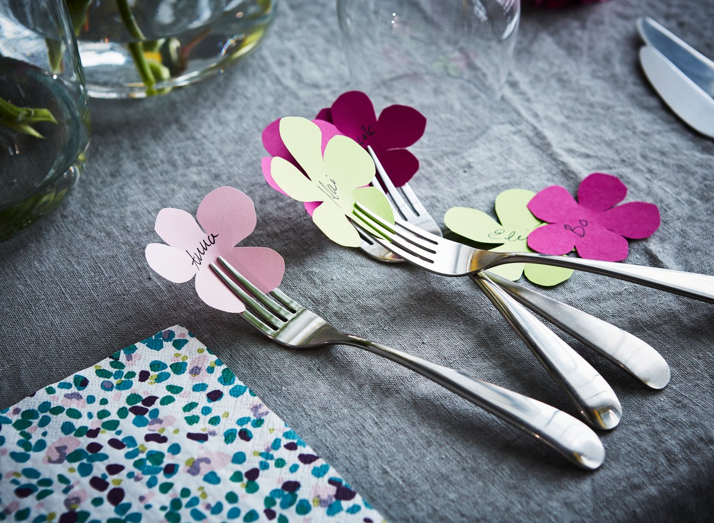 På et bord med en grå dug ligger gaffler med små blomsterformede kartonudklip, hvor der står et navn på.