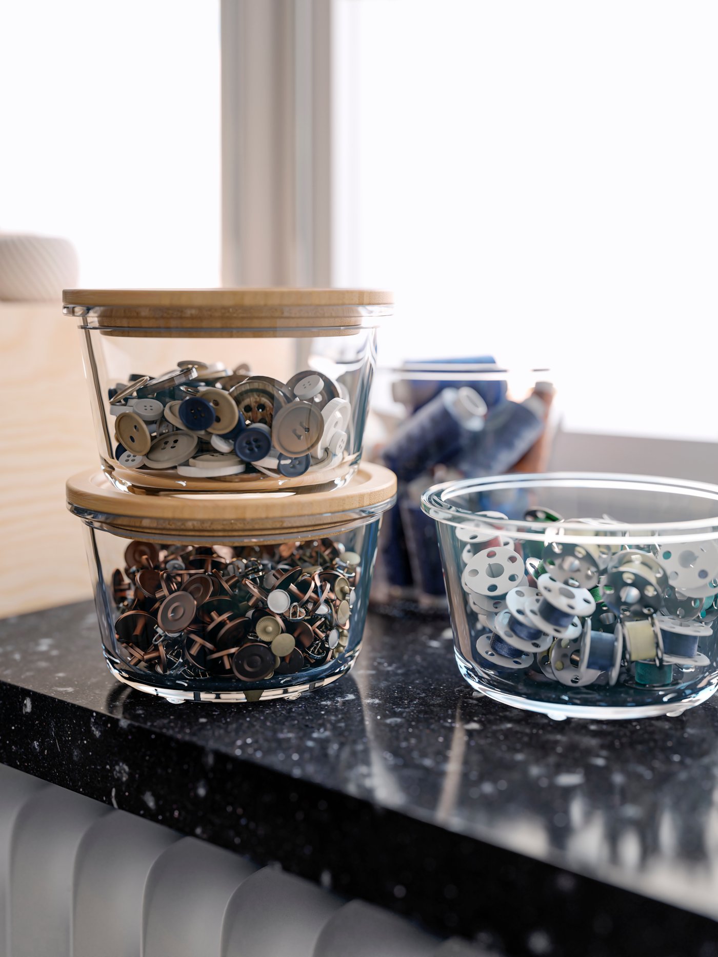 A windowsill with buttons, bobbins and other sewing accessories in IKEA 365+ glass food containers, some with bamboo lids.