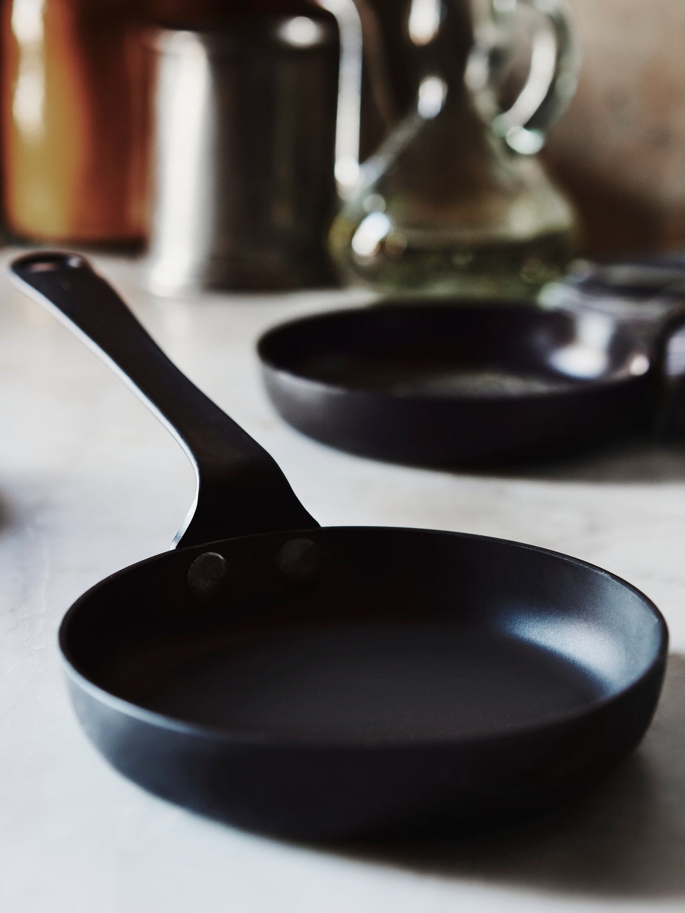 A black VARDAGEN frying pan placed on a grey marble countertop with a frying pan and kitchen accessories at the back.