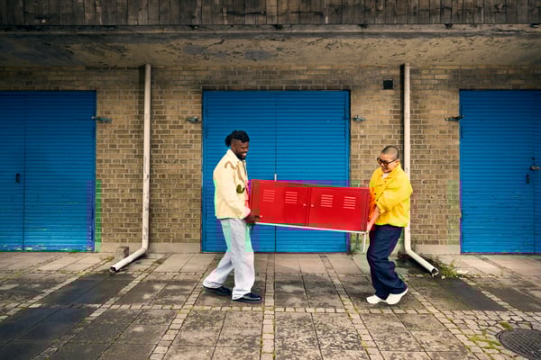 Dues persones transporten juntes un moble baix vermell davant d'uns garatges amb portes blaves. Escena urbana que reflecteix col·laboració i reutilització de mobles.