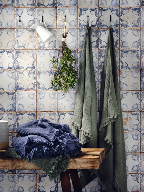 Bathroom area featuring two bath towels, a vase, and branches hanging from wall hooks. Under the vase is a bench with towels.