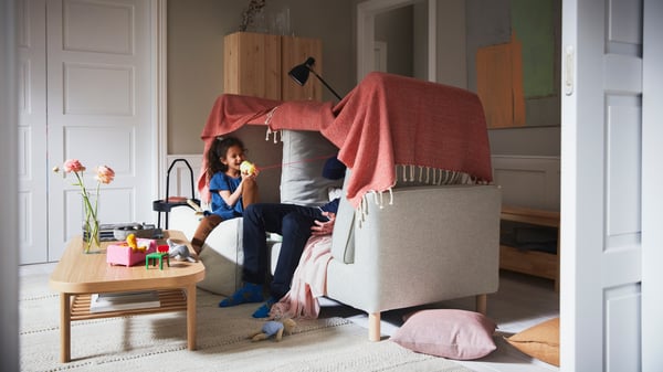Ett barn och en vuxen pratar i en trådtelefon i ett tält gjort av en beige PÄRUP soffa och en rödbrun/vit ULLTISTEL pläd.