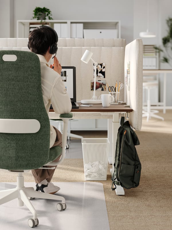 A man with a headset sits at a MITTZON desk with a MITTZON acoustic screen. A DRÖMSÄCK backpack hangs on a hook on the desk.