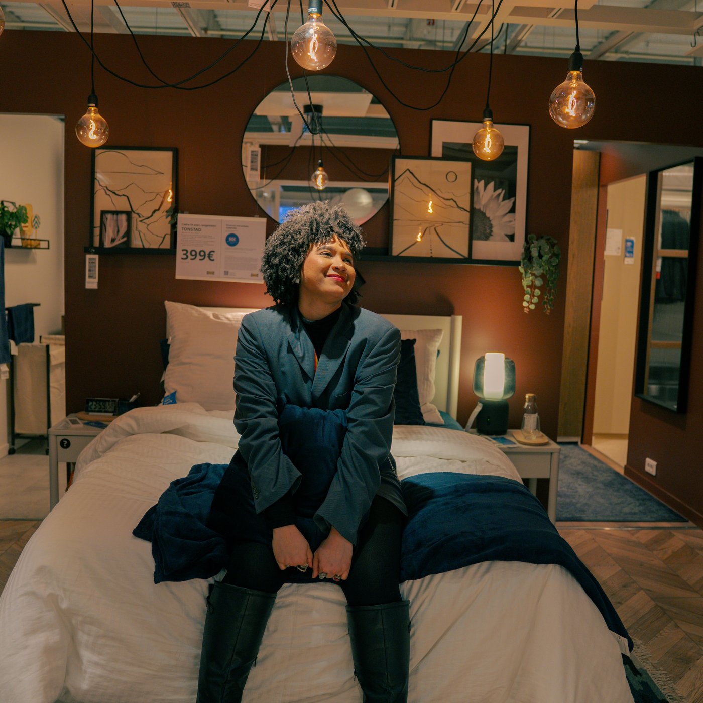 Shadia is seated on a bed, wearing a dark jacket and black boots, with curly hair and a relaxed posture, smiling gently.
