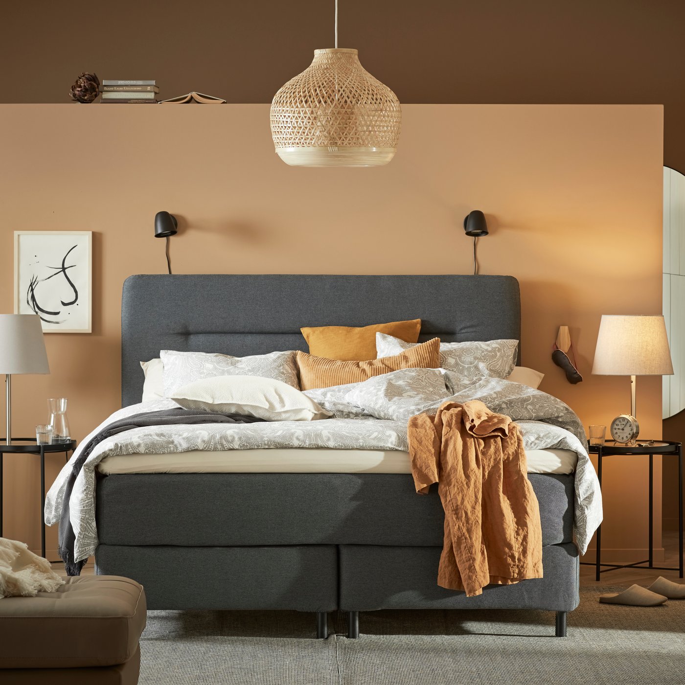 Een grote grijze boxspring in een kamer met warme kleuren als oranje en zandkleuren.