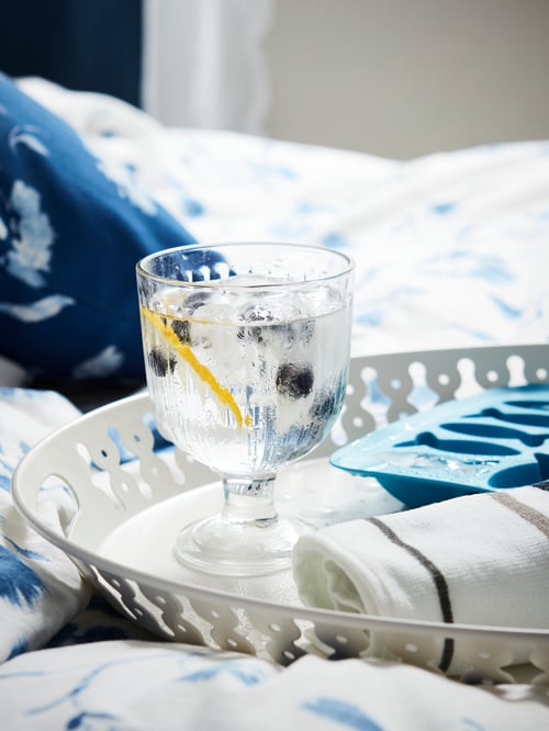 A white ROMANTISK tray with glass goblet and an ice cube tray on a bed with blue and white quilt cover and pillowcase.