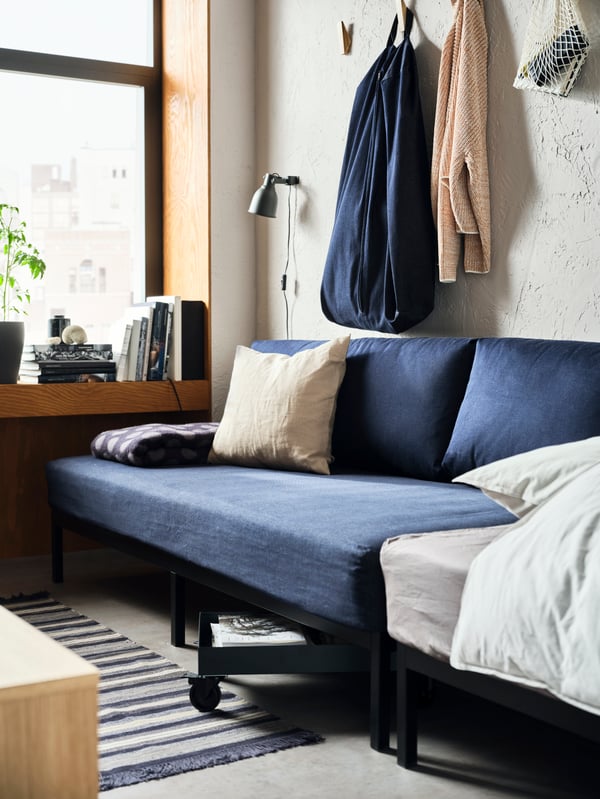 A dark blue RÅVAROR daybed is set against a concrete-style wall. A RÅVAROR storage bag has been hung above it.