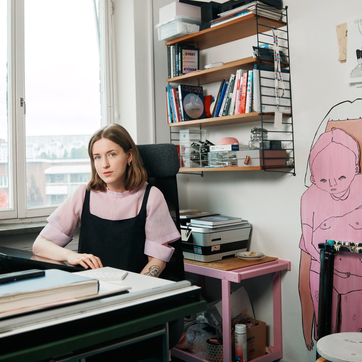 A woman is sitting behind a desk by a window. Behind her are shelves full of books and colourful cardboads cut in shapes of human.