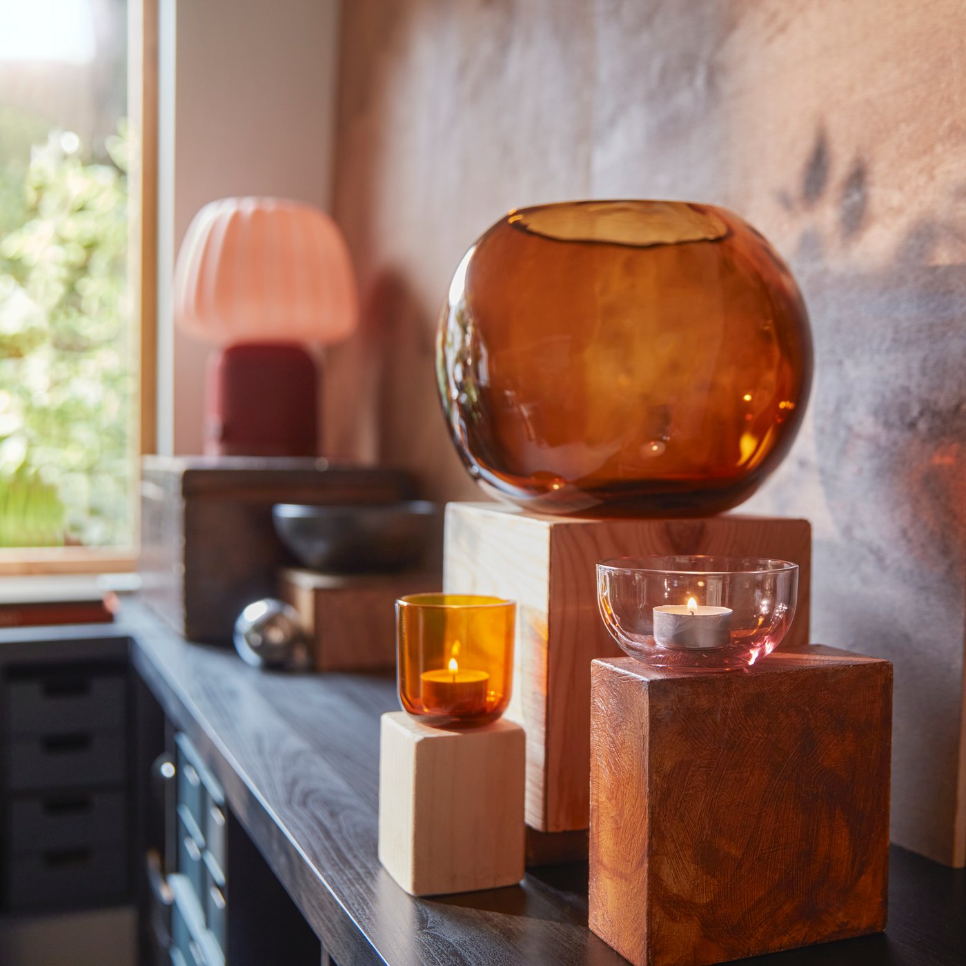 مجموعة من حاملات شموع صغيرة TALLTITA باللون الوردي/البني موضوعة على قوالب خشب ومحاطة ببعض أغراض الزينة.