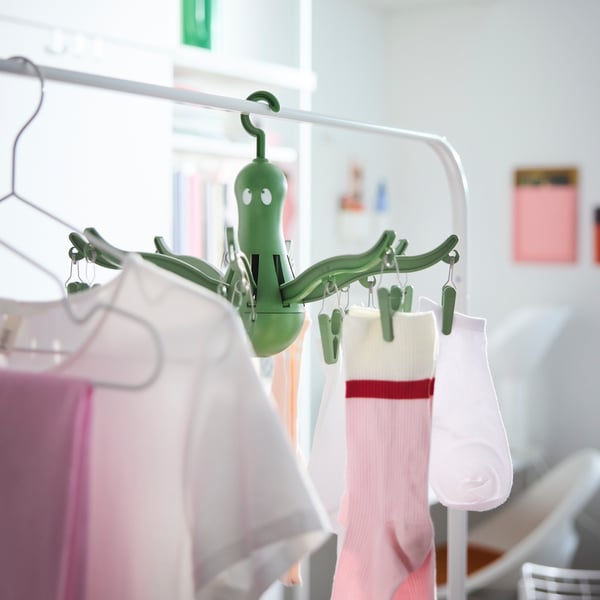 A green PRESSA hanging dryer with 16 clothes pegs hanging on a clothes rail in a white room, some pegs being used for socks.