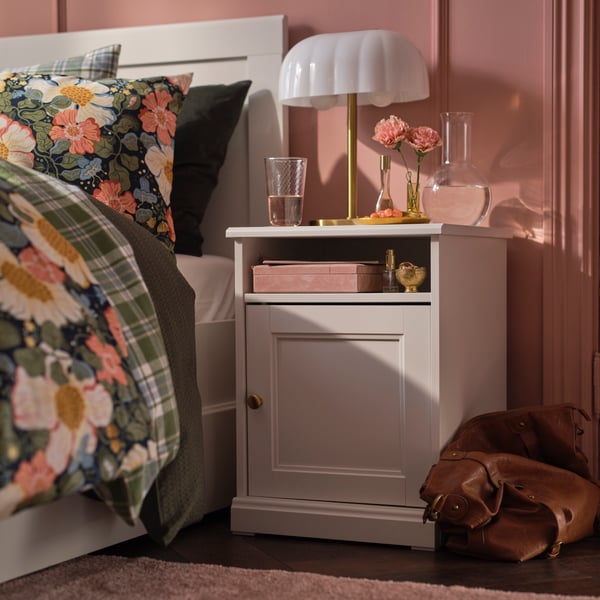 A FÅGELFJÄLLET side table in off-white with a white table lamp stands in a pink bedroom next to a bed with floral bedding.