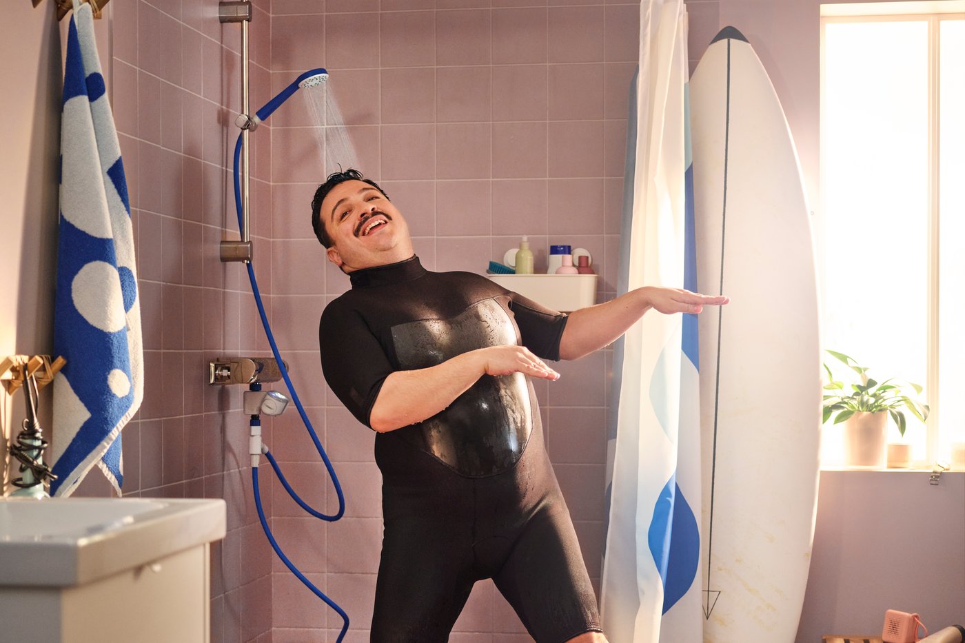 Un uomo sta facendo una doccia all'interno di un bagno, indossando una muta. Accanto alla tenda della doccia è appoggiata una tavola da surf.