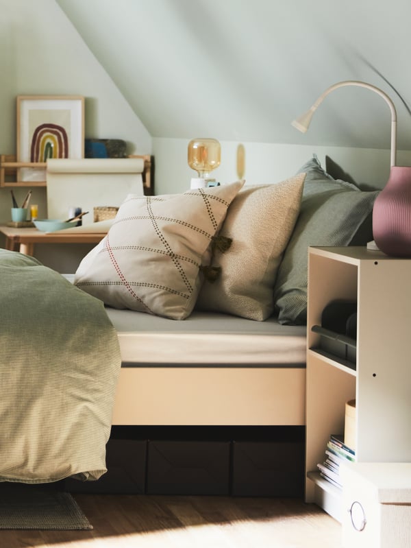 A light beige GURSKEN bed with various cushions by a small GURSKEN shelving unit with a white table lamp and pink vase.