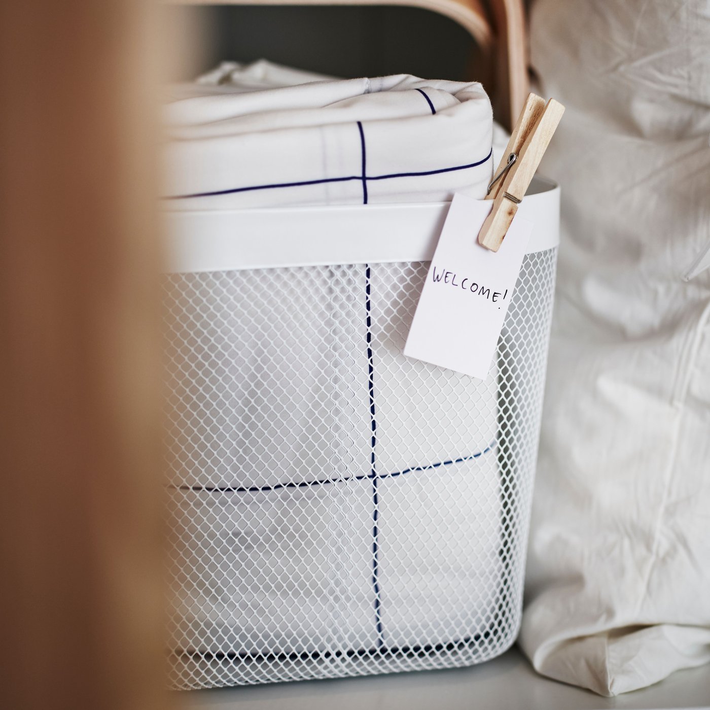 سلة RISATORP بيضاء عليها ملصق في الأمام على رف بها مفارش السرير BREDVECKLARE مزينة بنقش مربعات بالأبيض والأزرق مطوية.