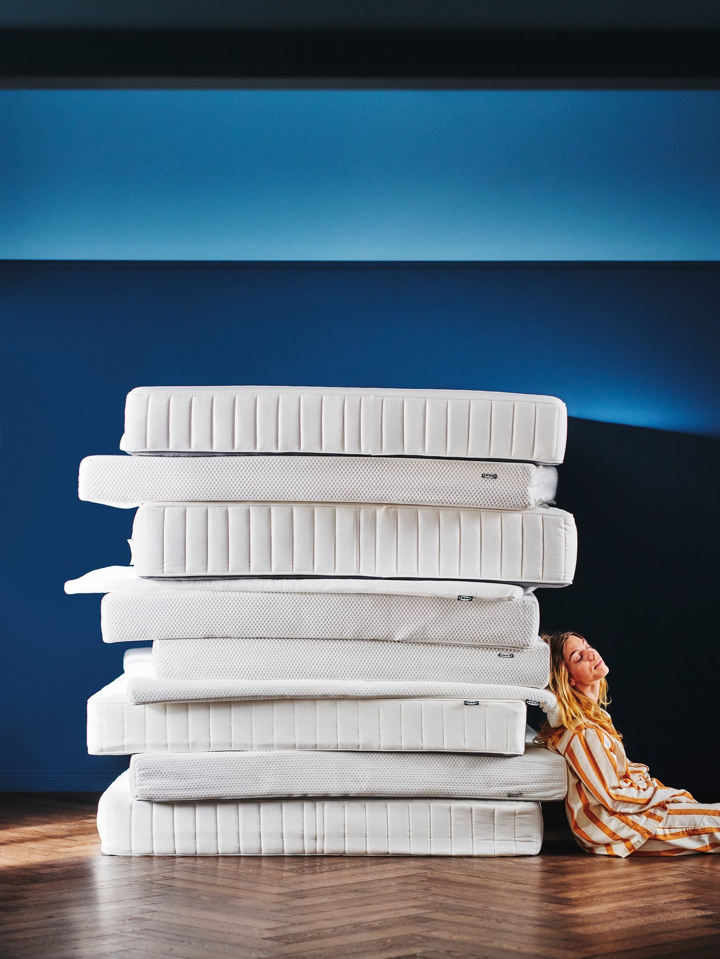 In a blue room, a person in striped pyjamas is sitting on the floor and leaning against a stack of IKEA mattresses.