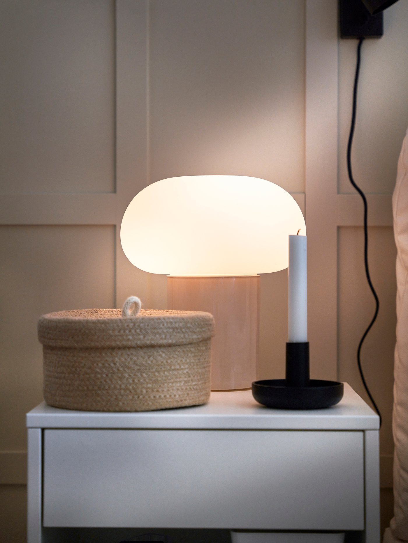 En liten bordslampa, rund korg med lock och en svart ljusstake med ett vitt ljus stående på ett vitt nattduksbord.