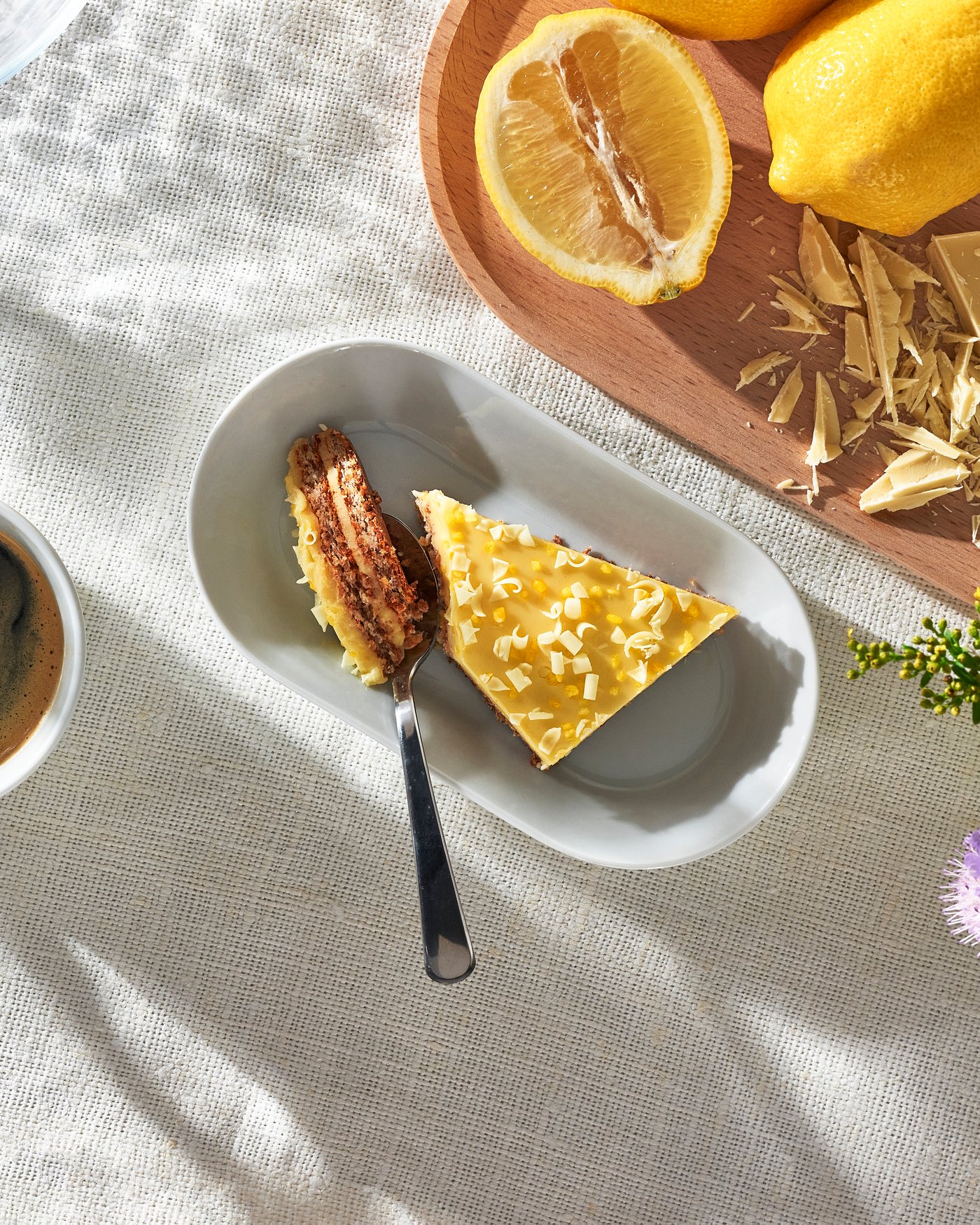 Auf einem weißen Teller liegt ein Stück Zitronenkuchen mit heller Glasur und feinen weißen Streuseln.
 Daneben stehen frische Zitronen und eine Tasse Kaffee auf einer hellen Stoffoberfläche.
