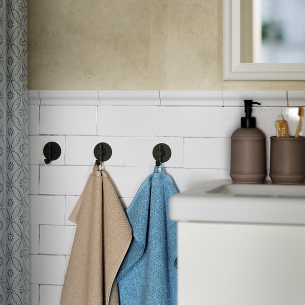 A beige GULVIAL guest towel hangs on one of three hooks on a tiled wall by a dark grey-blue towel and a bathroom wash-basin.