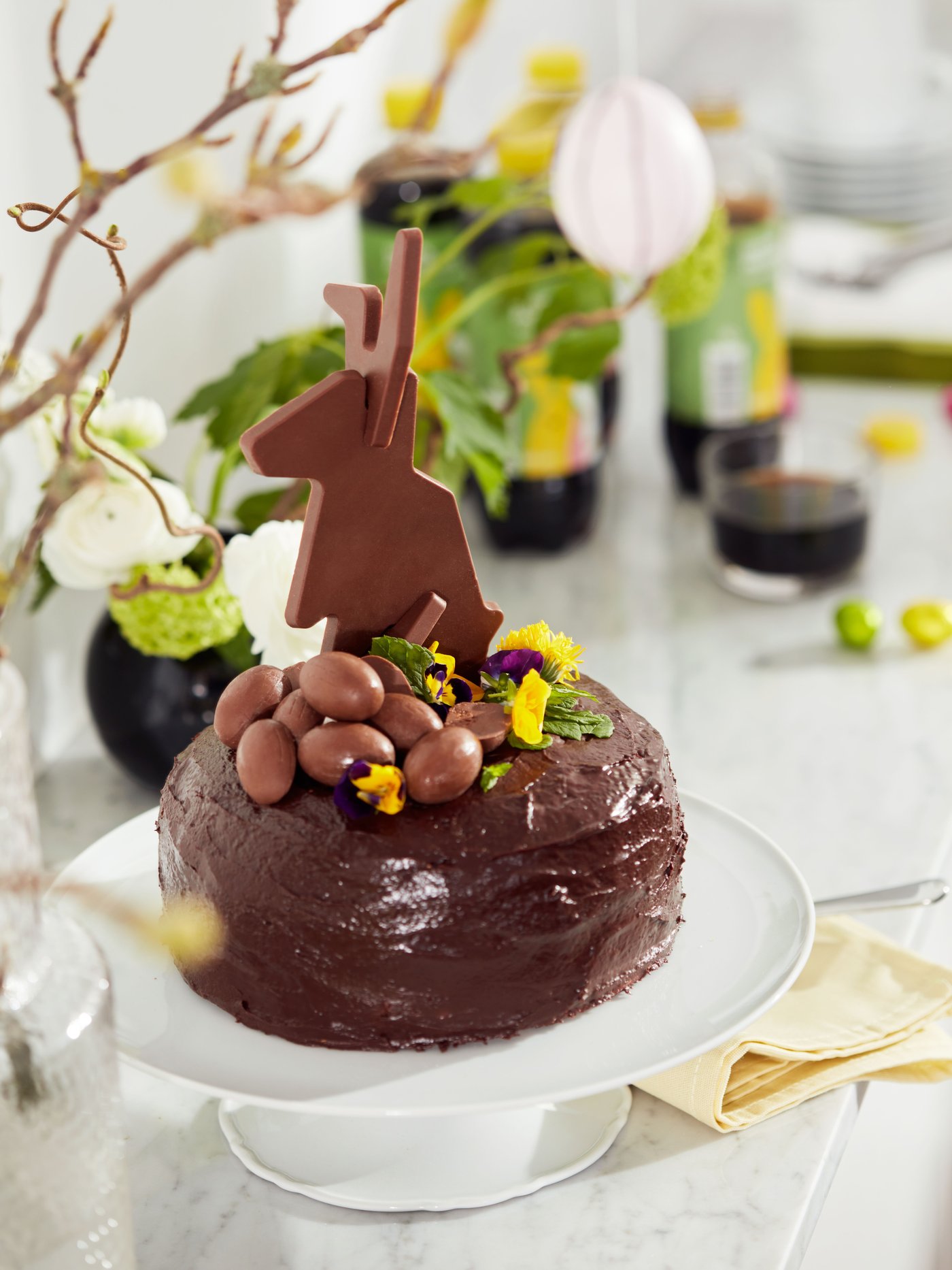 A chocolate cake with a chocolate Easter bunny on top on a white cake stand