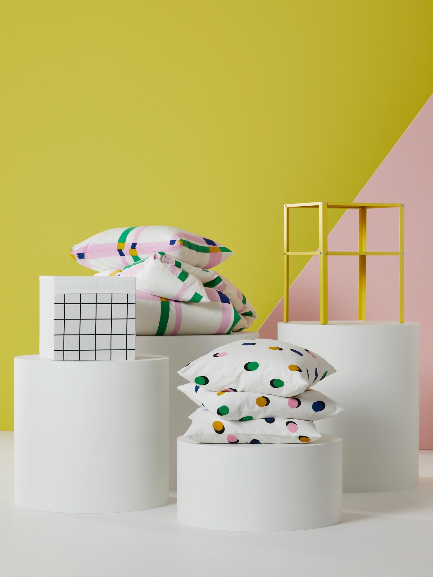 Tres cojines RUNDKRASSLIG en un podio. Detrás, también en podios, hay una mesa KNARREVIK, almohadas SYRABLADMAL y una caja DRÖNA.