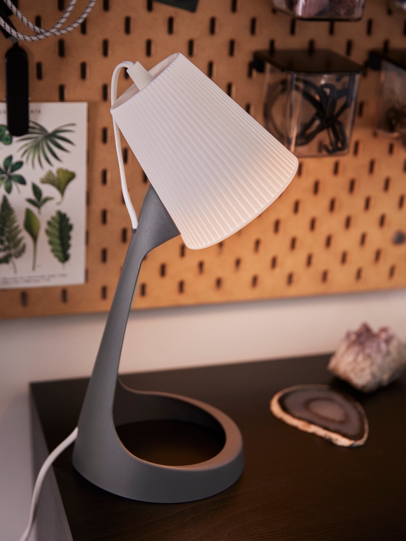 A lit white and dark grey SVALLET work lamp on a desk and a pegboard with a container with lid on the wall behind.