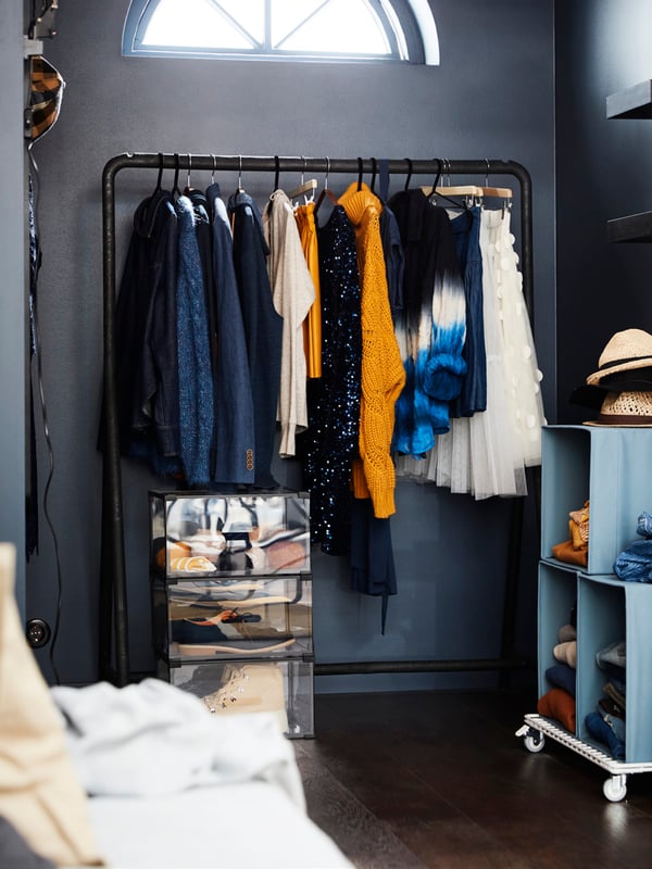A corner of a dark-wall room with a lunette window, with clothes hung on a TURBO clothes rack and folded in blue DRÖNA boxes.