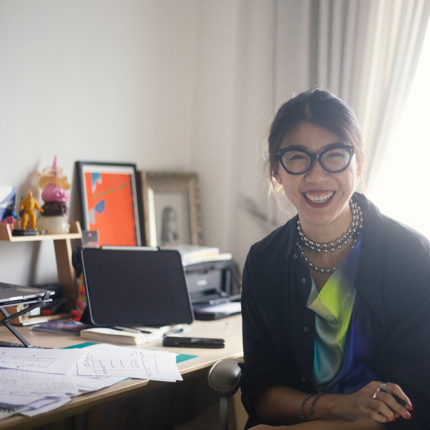 A smiling woman with glasses on is sitting by a desk full of papers and pencils. 