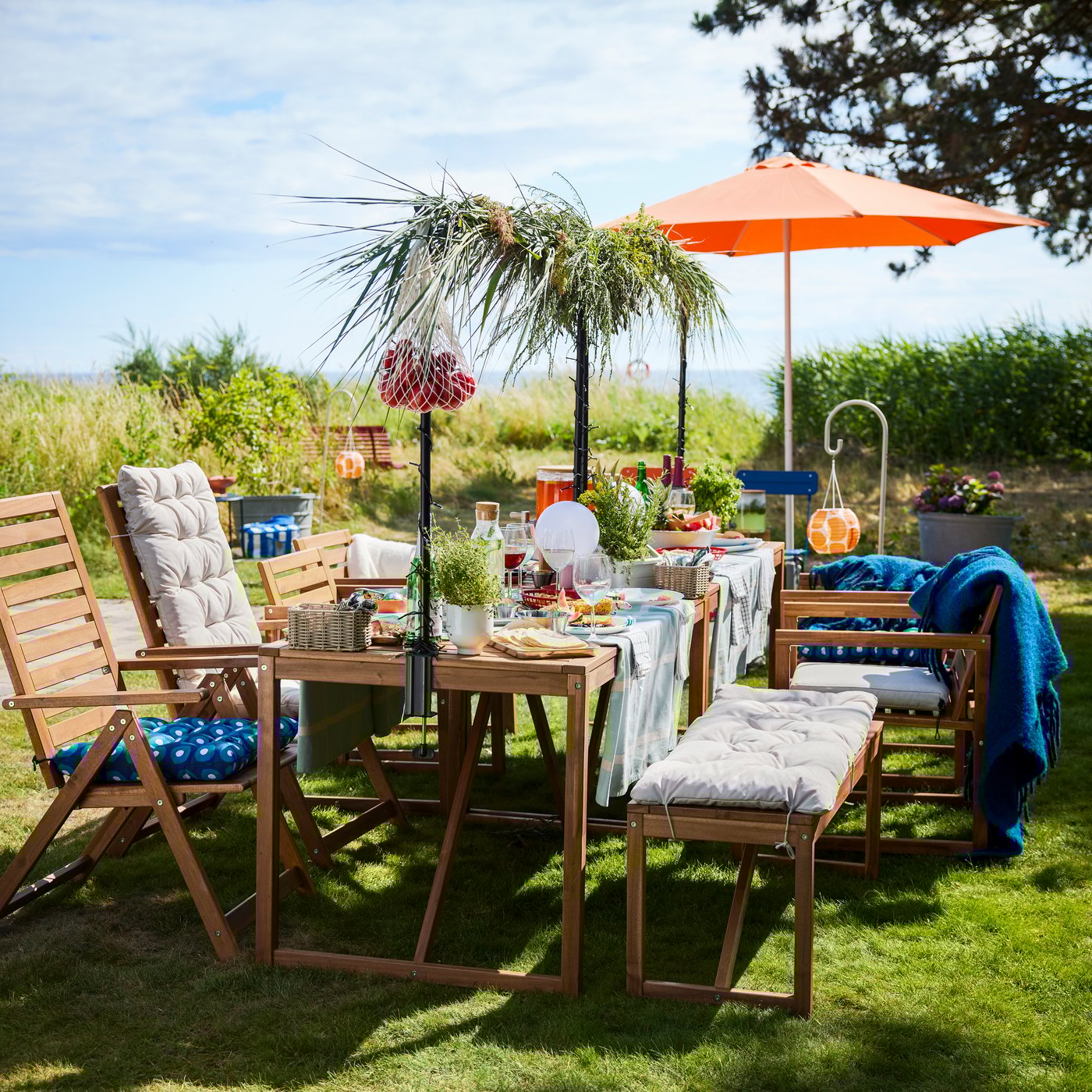 A garden setting with a light brown wooden NÄMMARÖ table with food and drinks on a sunny day. Chairs, a bench and a parasol.