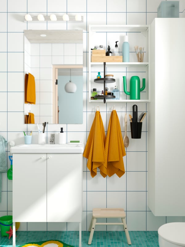 A white-tiled bathroom with a turquoise floor has a white ENHET base cabinet with legs and knobs and yellow towels on hooks.