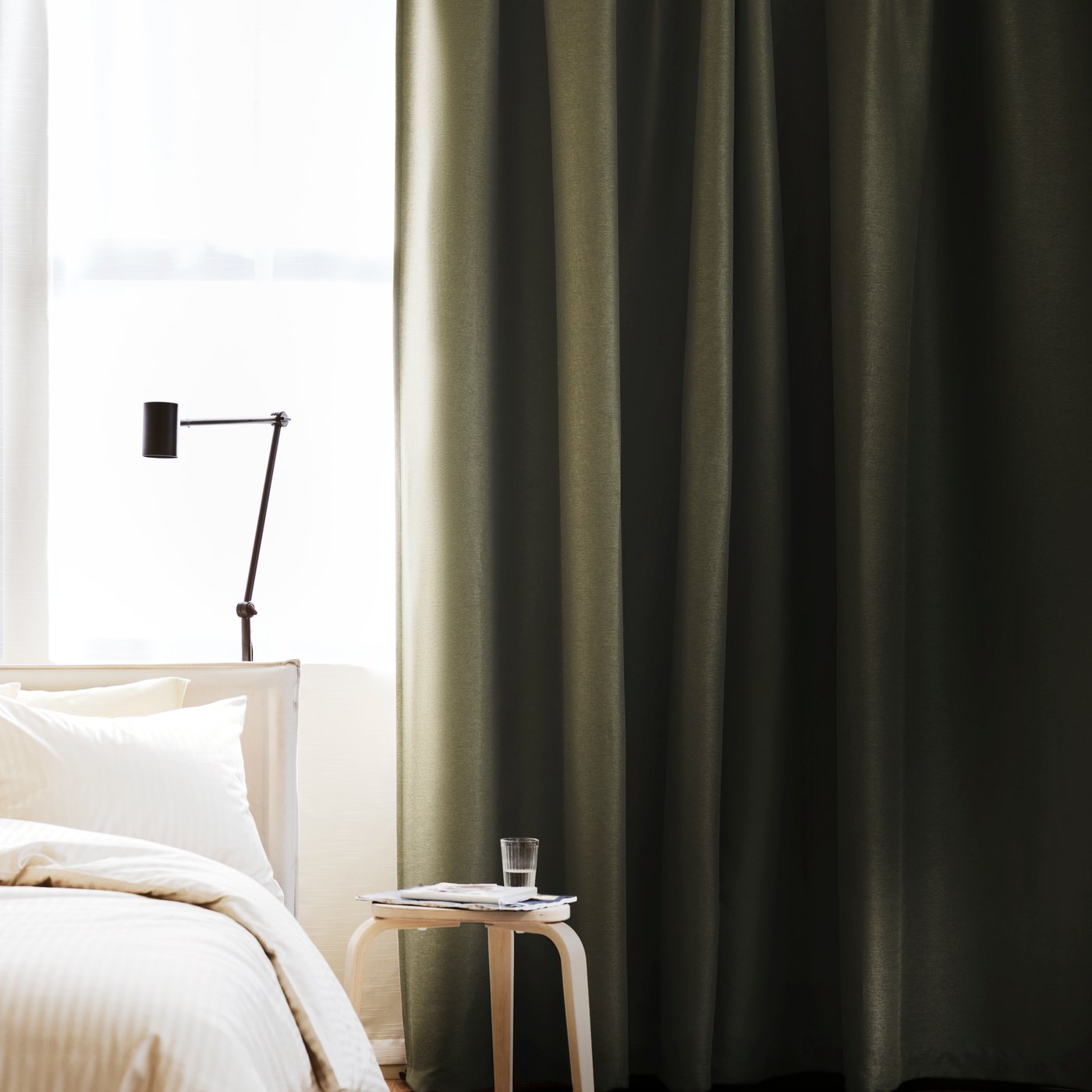 Sovrum med mörkgröna mörkläggningsgardiner som täcker fönstret, bredvid en bädd med ljust sänglinne och ett litet sidobord med glas vatten.
