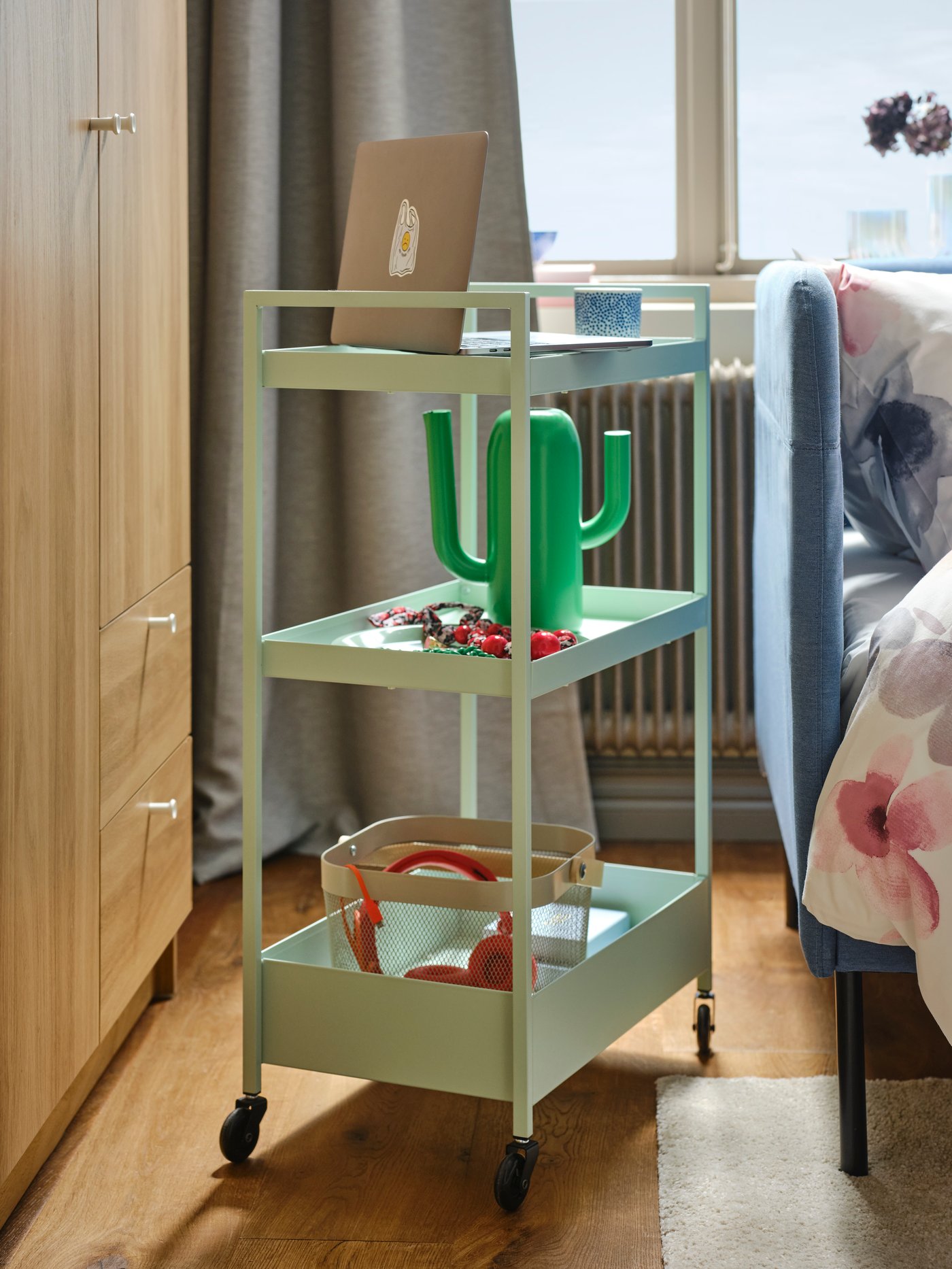 A pale green NISSAFORS trolley in a bright room with a laptop, an ÄRTBUSKE vase/watering can and a basket on its shelves.