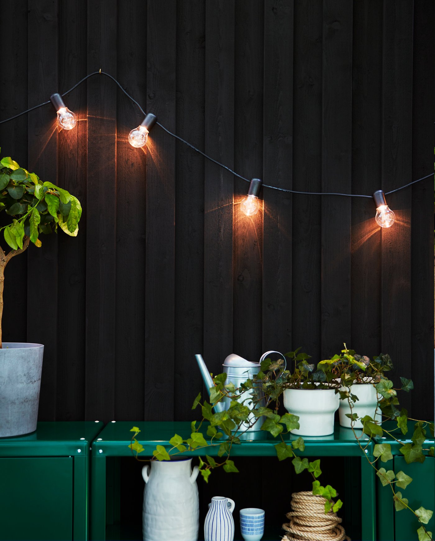 Guirnalda SVARTRÅ de 12 luces LED para exterior en color negro, instalada sobre una pared oscura y rodeada de plantas en una terraza.