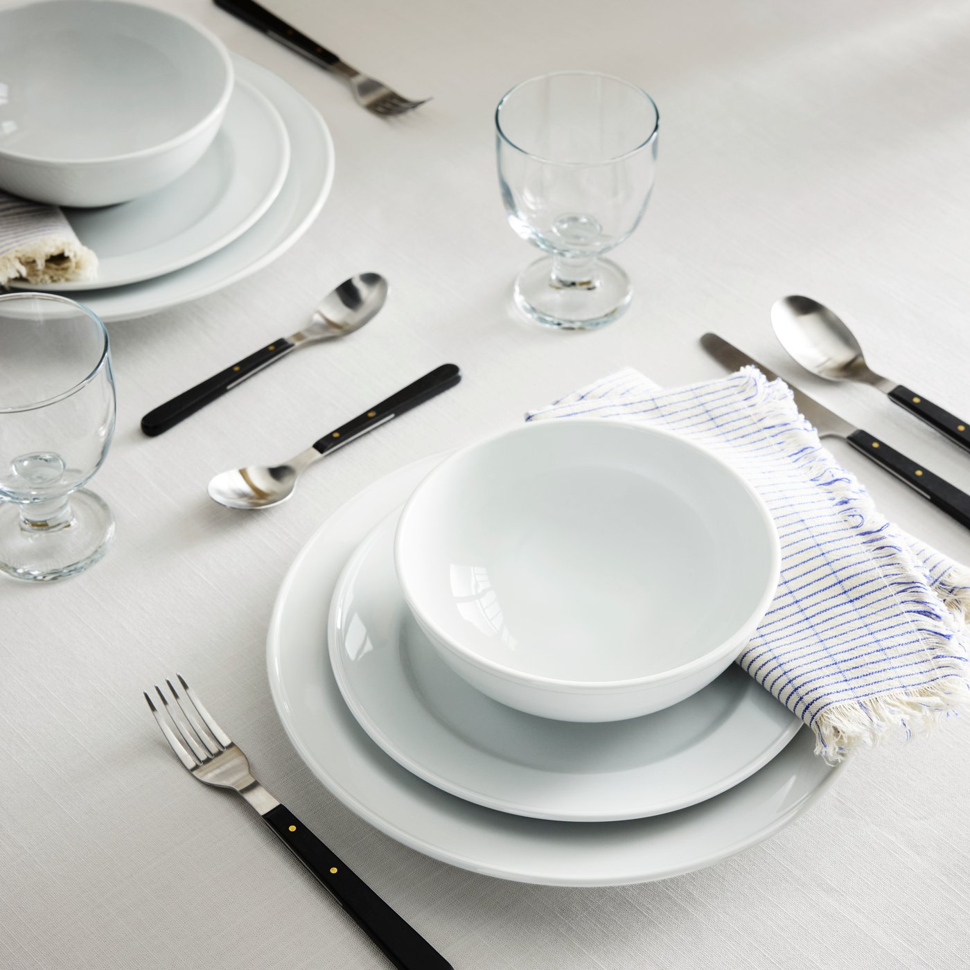 A table with a white tablecloth is set with VARDAGEN bowls, plates and side plates, DOFTSKÖLD cutlery and FRUKTDUVA goblet.