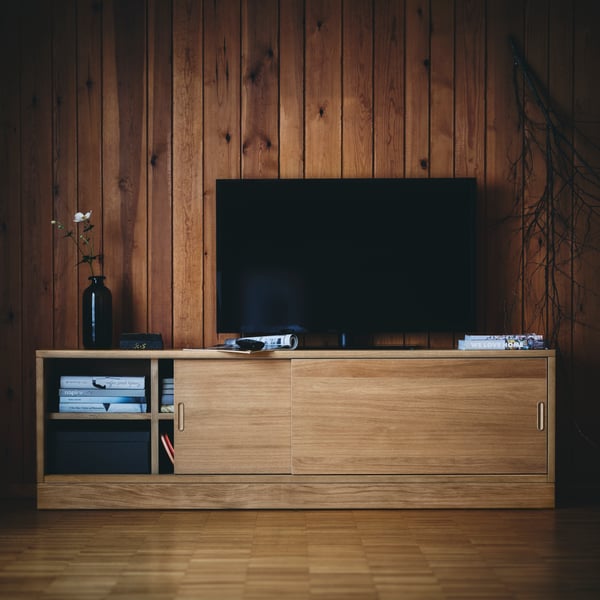 A TV and a vase with some flowers in it stand on top of an oak veneer TONSTAD TV bench with one door open.