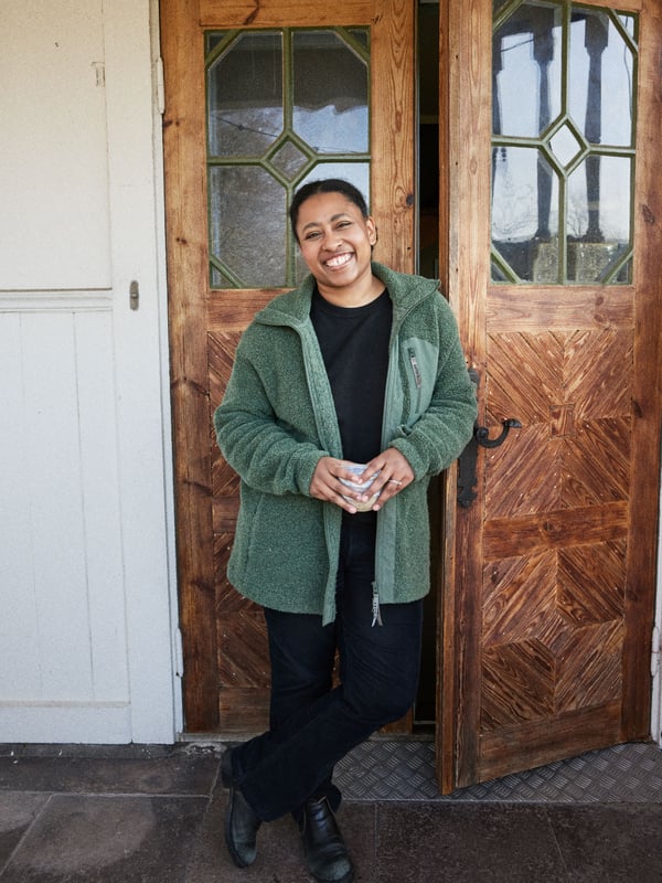 A woman standing with a white mug in her hands smiling with a brown door behind her.
