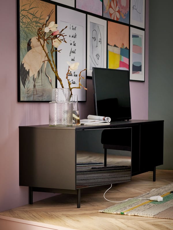 A RANNÄS TV bench with drawers with a vase and a TV on top of it stands against a wall which has a lot of BILD posters on it.