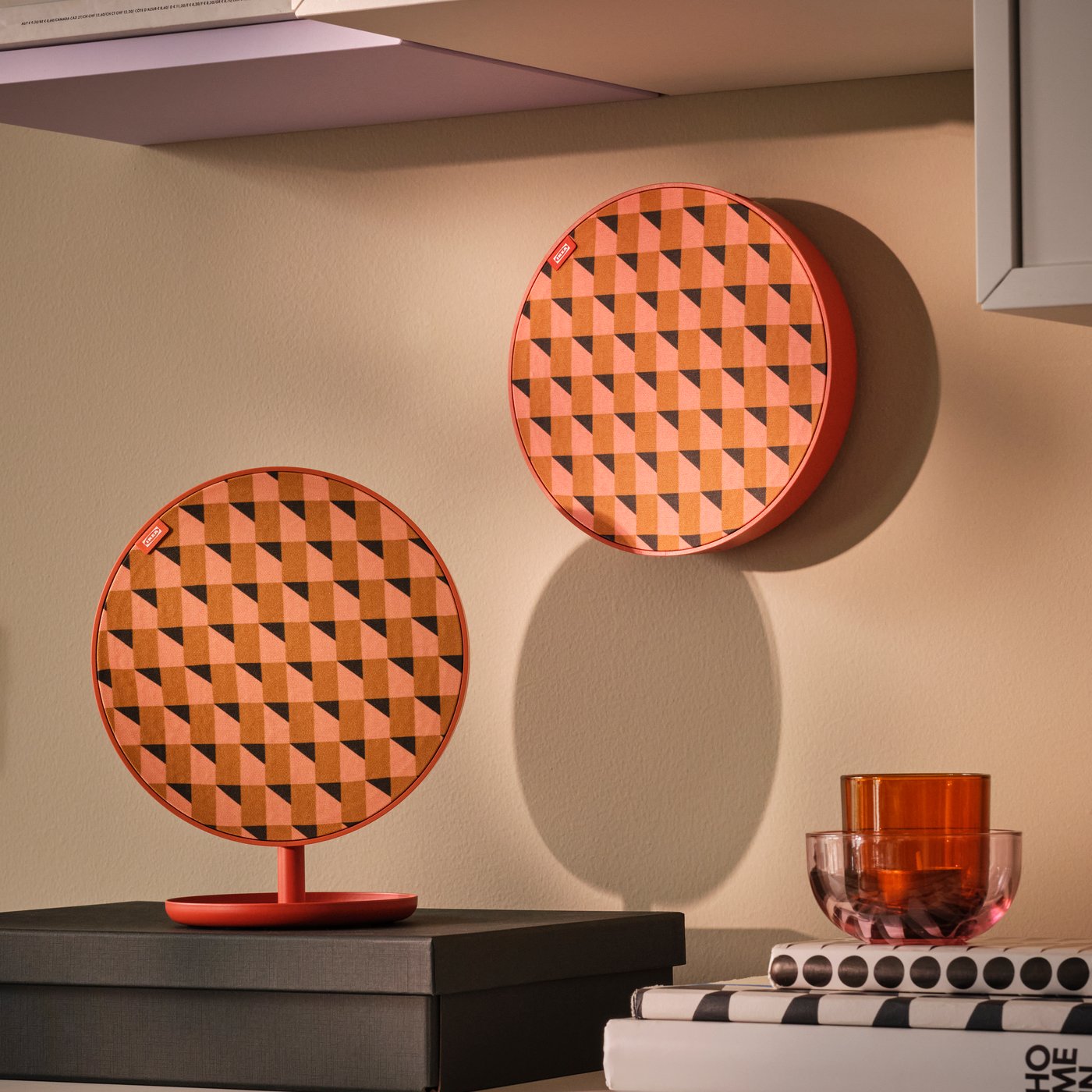 Due oggetti decorativi rotondi con un motivo geometrico arancione e nero, uno appoggiato su una base arancione e l'altro appeso alla parete, sono mostrati in un interno con un bicchiere arancione su una pila di libri.